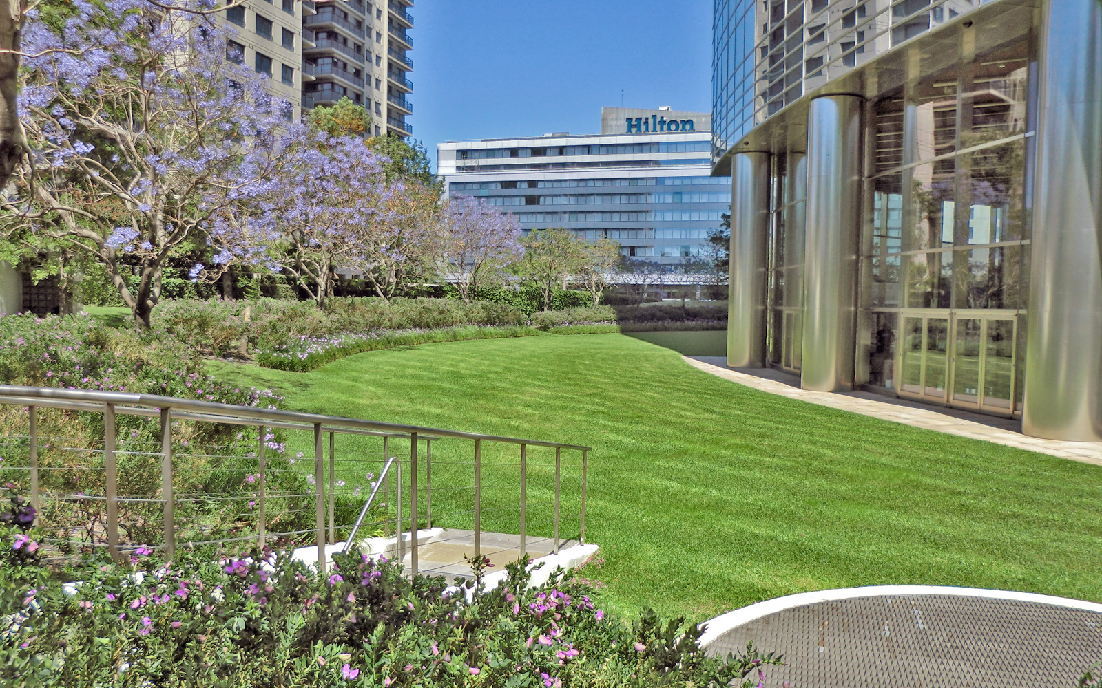 Parque con césped, árboles de Jacaranda en flor y entrada peatonal al aparcamiento subterráneo